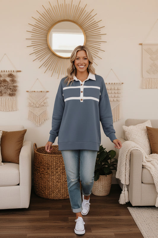 Woman standing in a livingroom wearing a blue long-sleeve shirt with white stripes and light blue jeans.